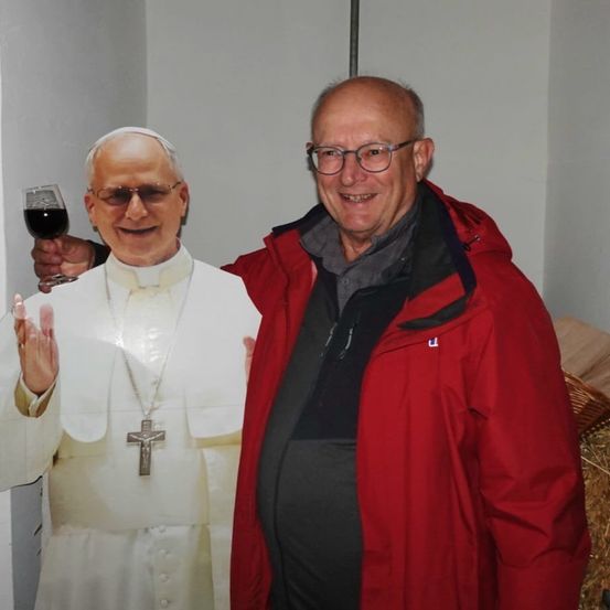 Ein Mann mit Brille und roter Jacke steht neben einem lächelnden Ausschnitt eines Papstes und hält ein Glas Wein in der Hand.