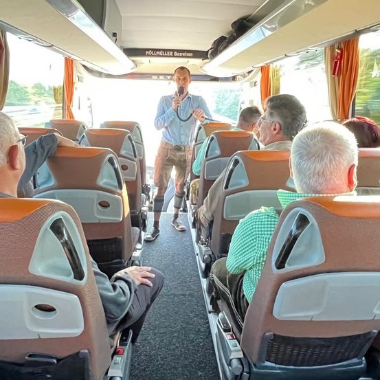 Ein Mann in einem hellblauen Hemd steht im Gang eines Busses, hält ein Mikrofon in der Hand und spricht die sitzenden Passagiere an. Der Bus ist voll mit älteren Erwachsenen, einige tragen Brillen, und das Innere hat orange Vorhänge und Gepäckträger über dem Kopf.