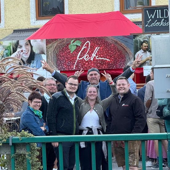 Eine Gruppe von Menschen steht unter einem roten Regenschirm. Die Frau in der Mitte lächelt. Sie posieren für ein Foto vor einem Schild, das Wild Lebküchner sagt.