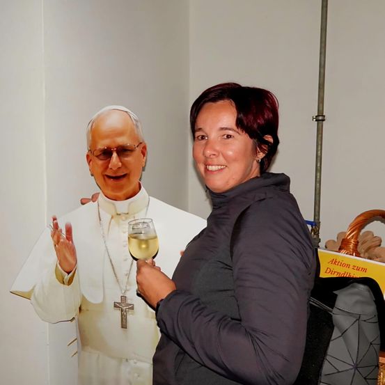 Eine lächelnde Frau hält ein Glas Wein in der Hand und steht neben einer Pappaufsteller eines lächelnden Papstes. Hinter der Frau ist ein Korb mit dem Text Aktion zum Dirndl zu sehen.