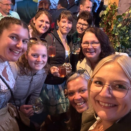 Eine Gruppe lächelnder Erwachsener, einige mit Brille, posiert mit Getränken bei einer Veranstaltung für ein Foto.