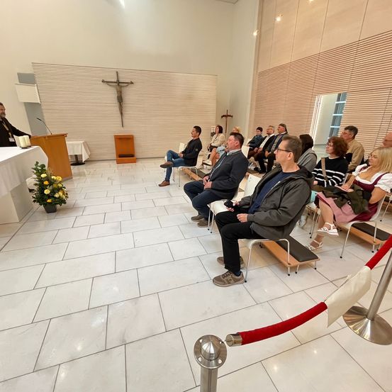 Eine Gruppe von Menschen sitzt in einer Kirche auf Stühlen und nimmt an einem Gottesdienst teil. Der Mann am Rednerpult spricht. An der Wand befindet sich ein Kreuz und auf dem Tisch steht ein Blumentopf.