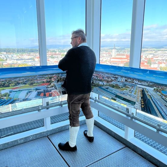 Ein Mann in traditioneller Kleidung steht auf einem Glasbalkon und schaut auf die Stadtlandschaft unten. Die Aussicht umfasst zahlreiche Gebäude und einen markanten Turm.