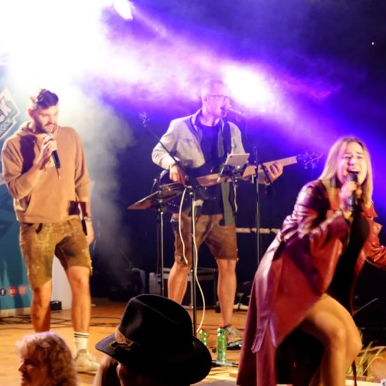 Drei Personen treten auf der Bühne mit lila und weißer Beleuchtung auf. Ein Mann singt und spielt Gitarre, ein anderer Mann singt und spielt Schlagzeug und eine Frau singt. Hinter ihnen schaut eine Menge zu.