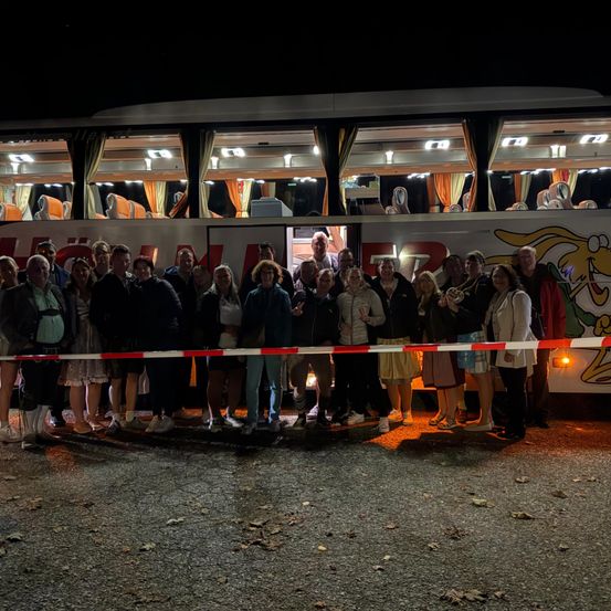 Eine Gruppe von Menschen steht bei Nacht vor einem Bus, einige Fenster sind beleuchtet und zeigen das Innere und Menschen darin.