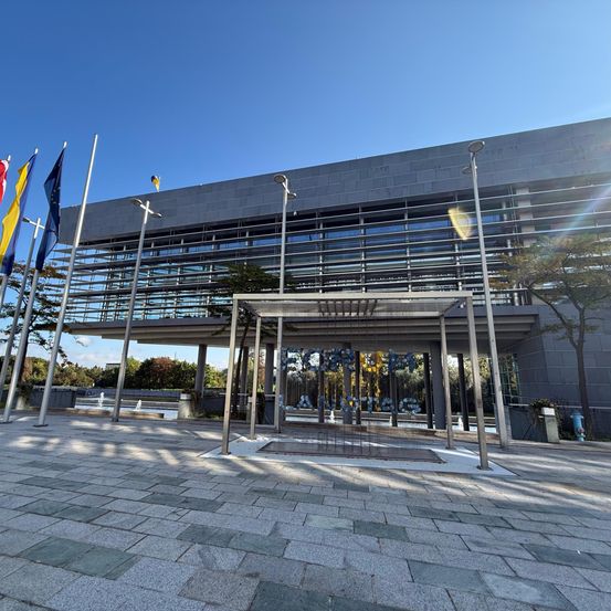 Ein modernes Gebäude mit Glaswänden und einem Eingangsbaldachin, umgeben von einer gepflasterten Fläche mit Fahnen und Bäumen.