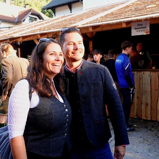 Ein Mann und eine Frau lächeln für ein Foto vor einem Holzstand mit einem Schild, das 'hier bestellen' steht. Hinter ihnen stehen andere Leute und unterhalten sich in der Nähe des Tresens.