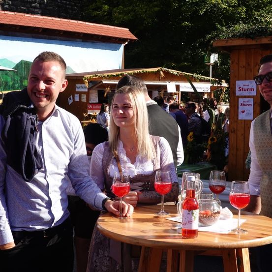 Zwei Personen stehen lächelnd vor einem Holztisch mit Gläsern roter Flüssigkeit. Dahinter unterhalten sich andere auf einem Festival mit Ständen und Schildern. Bäume und eine Wandmalerei sind im Hintergrund.