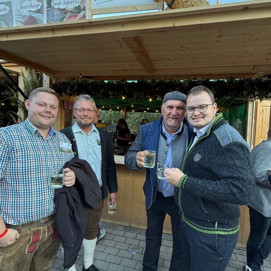 Vier Männer stehen vor einem Essensstand, Getränke in den Händen haltend. Sie lächeln und scheinen für ein Foto zu posieren. Hinter ihnen befindet sich eine Holzkonstruktion mit Text und Bildern darauf. Eine Person steht hinter dem Tresen, und einige Lampen sind an der Decke montiert.