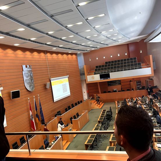 In einem modernen Konferenzsaal steht eine Person hinter einem Rednerpult, während eine andere Person eine Treppe hinaufgeht. An der Wand hängen Flaggen, und eine Projektionsleinwand zeigt Inhalte. Der Raum ist mit Stühlen und Tischen gefüllt, und eine Person schaut sich die Szene an.