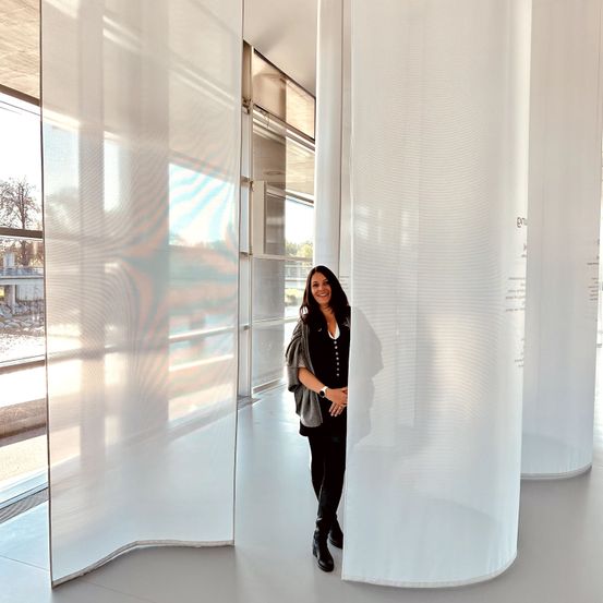 Eine Frau in einem schwarzen Outfit steht in einem architektonischen Raum mit weißen Stoffsäulen und großen Glasfenstern.