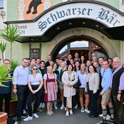 Eine Gruppe von Menschen steht vor einem Gebäude mit dem Schild 'Schwarzer Bar' über dem Eingang. Einige tragen Brillen und Schuhe. Pflanzen und Lampen sind in der Nähe.