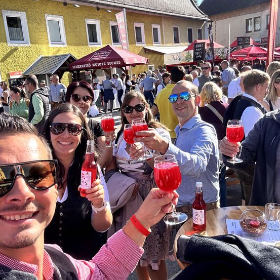 Eine Gruppe von Menschen mit Sonnenbrillen steht draußen, hält Getränke und lächelt. Sie sind um einen Tisch versammelt, auf dem eine Flasche und Gläser stehen. Im Hintergrund ist eine geschäftige Straße mit Zelten und Menschen zu sehen.