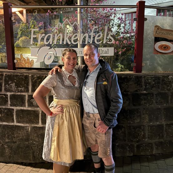 Ein Mann und eine Frau stehen nebeneinander vor einer Mauer und lächeln für ein Foto. Hinter ihnen befindet sich eine Glaswand mit dem Schild 'Frankenfels'.