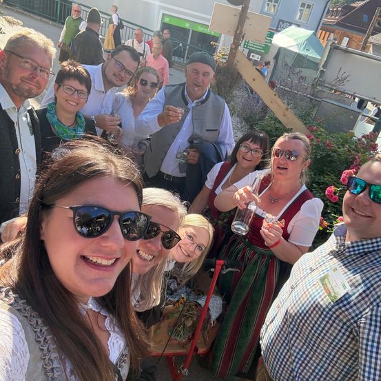 Eine Gruppe von Menschen in traditioneller deutscher Kleidung posiert für ein Foto, lächelt und hält Getränke in den Händen. Der Hintergrund zeigt eine festliche Umgebung mit Pflanzen und Gebäuden.