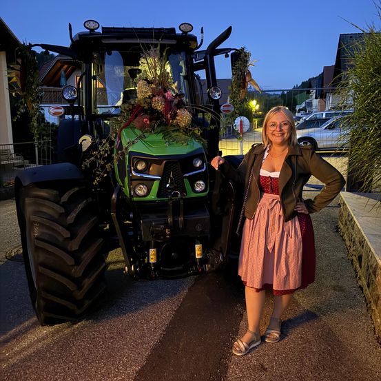 Eine Frau steht neben einem grünen Traktor, der mit Blumen geschmückt ist, und lächelt vor einem Haus in der Dämmerung.