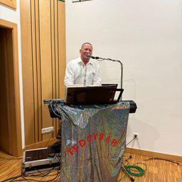 Ein Mann steht an einem Podium mit Laptop, Mikrofon und einem Tuch mit dem Wort Teddybar. Er lächelt und scheint eine Vorstellung zu machen.