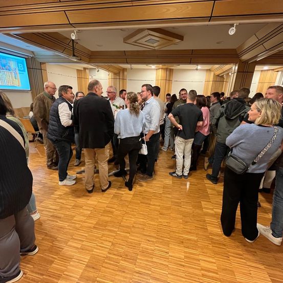 Eine Gruppe von Menschen steht und unterhält sich in einem Konferenzsaal. Einige tragen Brillen und andere haben Handtaschen. Eine Projektionsleinwand ist an der Wand montiert.