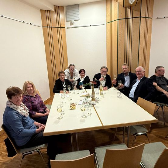 Eine Gruppe von Menschen sitzt um einen Tisch in einem Raum und hält Weinglaser in der Hand und lächelt. Sie scheinen ihre Zeit zu genießen.