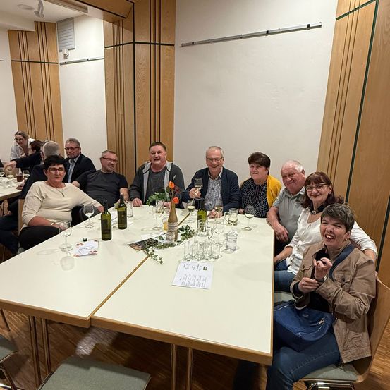 Eine Gruppe von Menschen lächelt und sitzt um einen Tisch mit Wein glasses, Flaschen und einem Papiermenü. Sie befinden sich in einem Restaurant mit Holzwänden.