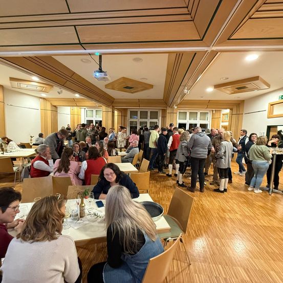 Ein Veranstaltungsraum mit Holzboden, in dem sich eine große Gruppe von Menschen versammelt hat. Einige stehen und unterhalten sich, während andere an Tischen sitzen. Der Raum hat Holzwände und Decke, und eine Projektor hängt an der Decke.