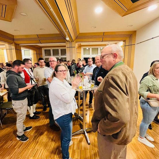 Eine Gruppe von Menschen in einem Raum mit Holzböden, einige stehen, andere sitzen und wieder andere unterhalten sich miteinander. Eine Frau klatscht und eine Tisch mit Getränken steht in der Mitte.