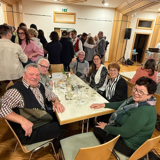 Eine Gruppe von Menschen sitzt an einem Tisch in einem Raum, einige lächeln und halten Weingläser. Es gibt Flaschen und Weingläser auf dem Tisch. Der Raum hat Holzwände und einen Lautsprecher auf der rechten Seite.