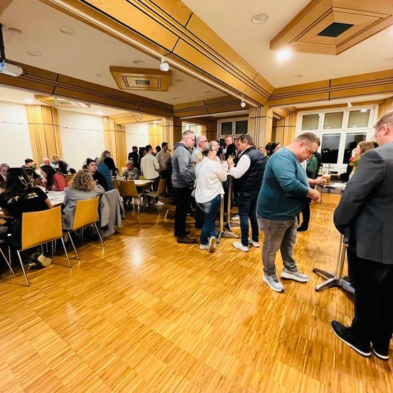 Eine Gruppe von Menschen steht in einem Speisesaal. Einige sitzen auf Stühlen, während andere in der Nähe von Tischen stehen. Eine Person hält ein Glas Bier. Der Raum hat Holzböden und Wände.