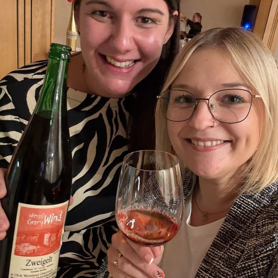 Zwei Frauen lächeln und halten eine Flasche Wein und ein Weinglas, eine trägt eine Brille und die andere hält die Flasche.