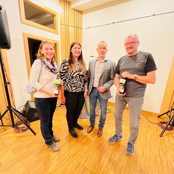 Vier Personen stehen in einem Raum mit Holzboden und halten Flaschen. Zwei Frauen und zwei Männer, alle lächeln. Hinter ihnen befinden sich Lautsprecher und eine weiße Wand.