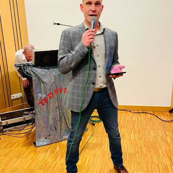 Ein Mann spricht in ein Mikrofon bei einer TEDx-Veranstaltung. Eine Person hinter ihm repariert Kabel. Der Hintergrund hat 'TEDx' darauf geschrieben.