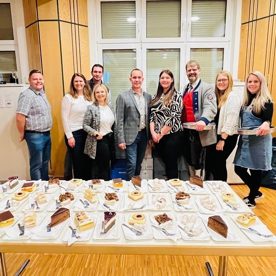 Eine Gruppe von Menschen posiert für ein Foto in einem Raum mit einem Tisch voller Desserts. Sie alle lächeln.