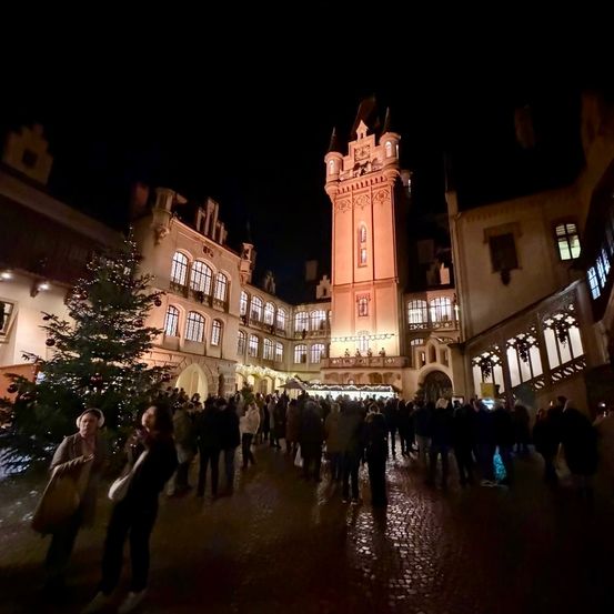 Ein überfüllter Stadtplatz bei Nacht mit einem hohen Turm, einem großen Weihnachtsbaum und herumlaufenden Menschen. Die Gebäude sind beleuchtet und es gibt viele Lichter.