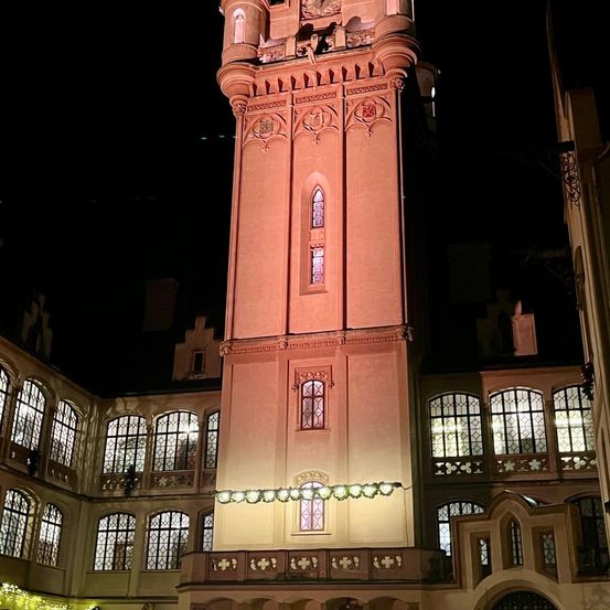 Ein hoher Turm im gotischen Stil, der nachts hell beleuchtet ist und vor einem Gebäude mit vielen Bogenfenstern steht.