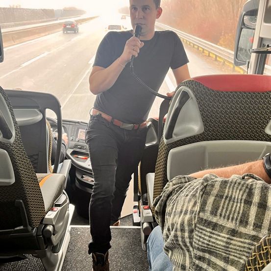 Ein Mann in einem Bus hält ein Mikrofon, möglicherweise macht er eine Ansage. Er steht neben dem Fahrer, und zwei Passagiere sitzen in der Nähe. Draußen fahren Autos auf einer Autobahn.