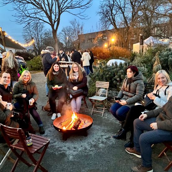 Eine Gruppe von Menschen sitzt bei Dämmerung um ein Feuer herum, einige halten Getränke und andere essen. Sie sind von Stühlen umgeben und ein Gebäude im Hintergrund.