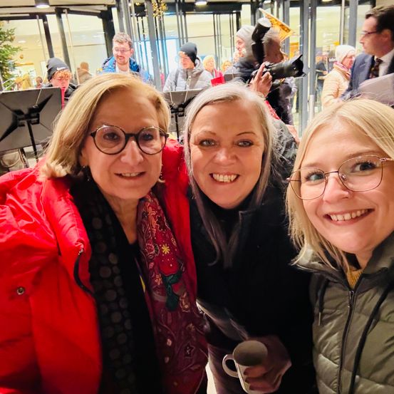 Drei Frauen in Mänteln lächeln für ein Foto mit einer Tasse im Vordergrund. Hinter ihnen spielen Leute Instrumente, machen Fotos und ein Mann spricht.