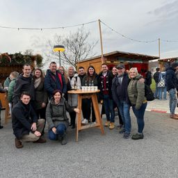 Eine Gruppe von Menschen posiert für ein Foto um einen kleinen Tisch mit Flaschen, vor dem Hintergrund von Winterdekorationen und kleinen Hütten.