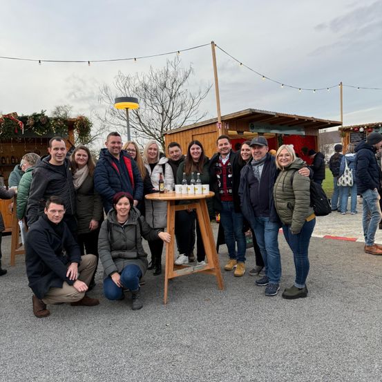 Eine Gruppe von Menschen posiert für ein Foto um einen kleinen Tisch mit Flaschen, vor dem Hintergrund von Winterdekorationen und kleinen Hütten.