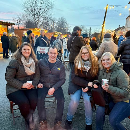 Vier Personen sitzen zusammen auf einem Markt im Freien und lächeln für die Kamera. Sie halten Tassen in der Hand, und die Umgebung umfasst Stände und Menschen in Winterkleidung.