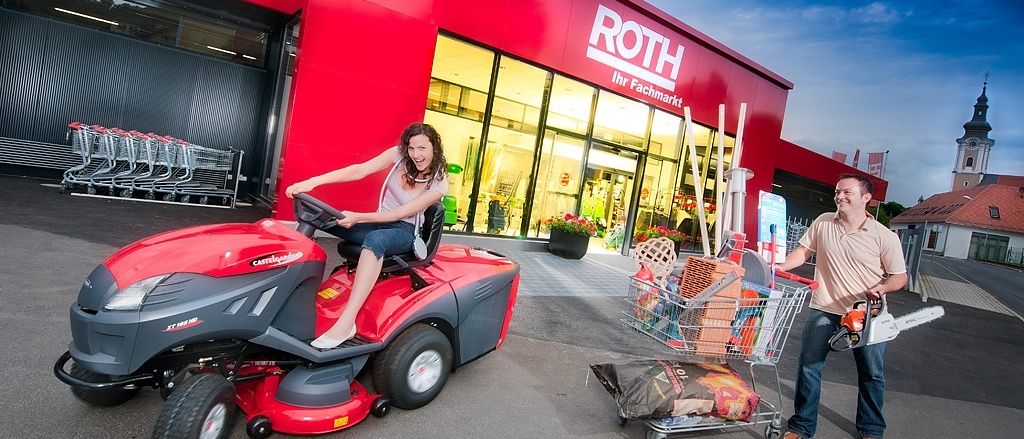 Eine Frau fährt einen roten Rasenmäher vor einem Geschäft mit dem Schild Roth. Ein Mann schiebt einen Einkaufswagen mit Einkäufen.