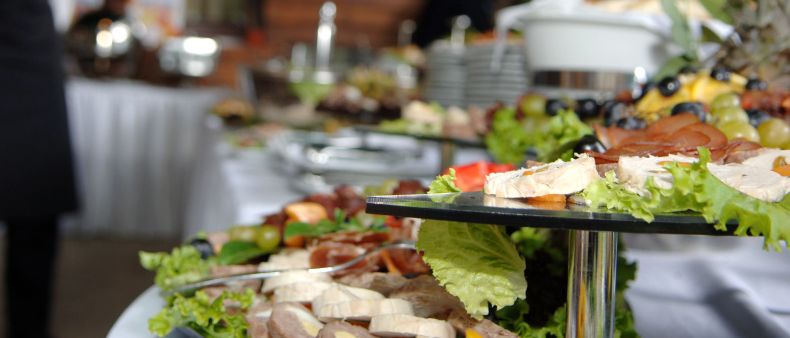 Ein Catering-Event mit mehreren Tabletts mit Essen, einschließlich Fleisch, Gemüse und Oliven, auf einem Tisch angeordnet, mit einer verschwommenen Person im Hintergrund.