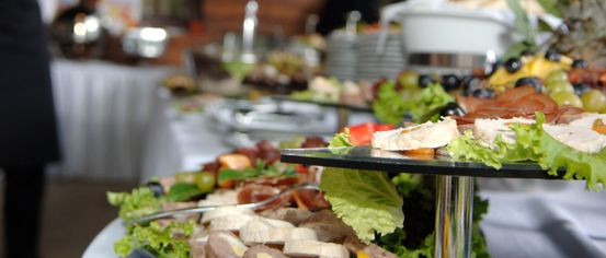 Ein Catering-Event mit mehreren Tabletts mit Essen, einschließlich Fleisch, Gemüse und Oliven, auf einem Tisch angeordnet, mit einer verschwommenen Person im Hintergrund.