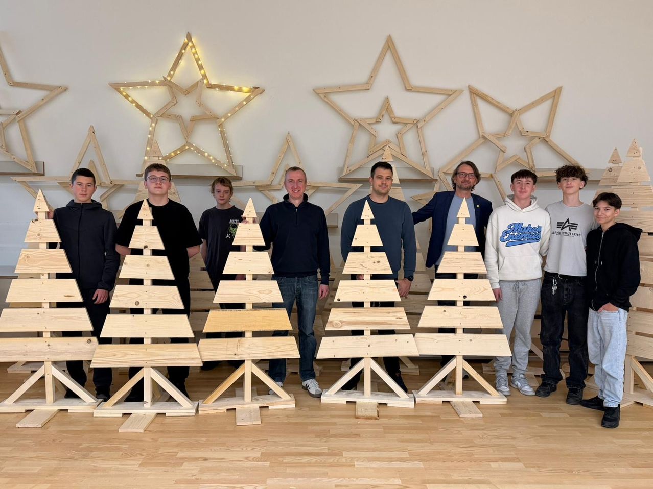 Eine Gruppe junger Männer und Erwachsener steht neben Holzweihnachtsbäumen und Sternendekorationen. Sie alle lächeln und posieren für ein Foto.