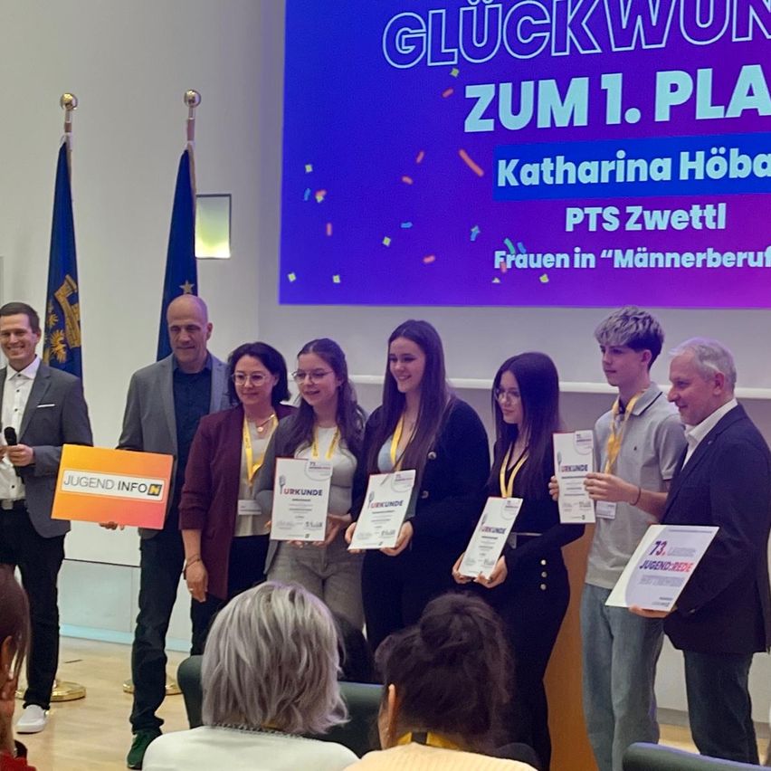 A group of people stand holding certificates. The screen behind them displays 'Congratulations to the 1st Place' and 'Katharina Höba, PTS Zwettl, Women in Male-Dominated Professions.' Three men and three women, some holding certificates, are standing in front of a podium.