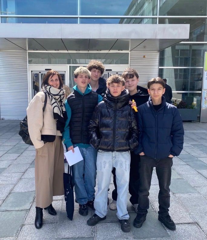 A group of people stand outside a modern building with glass walls. A woman in a beige coat holds a paper, and a boy in a black jacket poses. Other boys, one holding a yellow object, look in different directions.