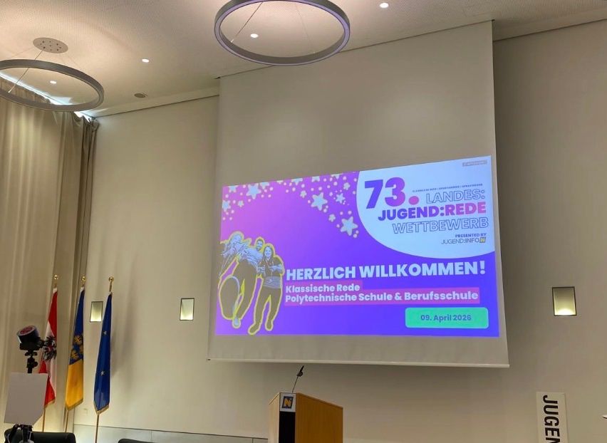 A conference room with a screen displaying the 73rd Landes Jugend Rede Wettbewerb event. Flags from various countries are on display.