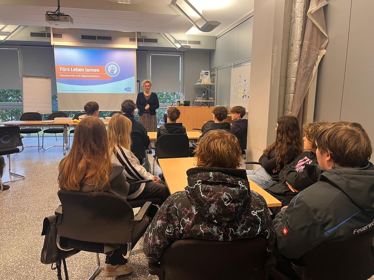 A woman stands in front of a classroom, presenting a lecture on a projector screen. Several students sit in chairs, listening attentively. The room is well-lit with natural light from the windows.