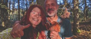 Ein Mann und eine Frau stehen in einem Wald, lächeln und posieren mit einem Pilz. Der Mann gibt einen Daumen hoch.