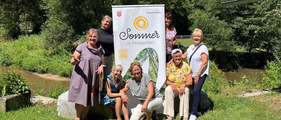 Fünf Frauen posieren vor einem Banner mit der Aufschrift 'Sommer in Pregarten'. Sie sitzen und stehen auf einem Rasen mit einem Fluss und Bäumen im Hintergrund.